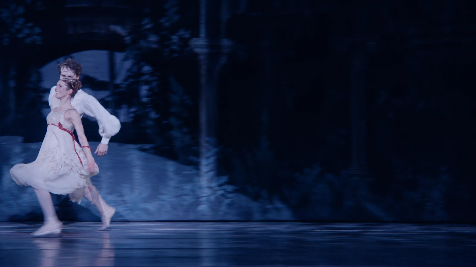 Two ballet dancers crossing a dark blue stage